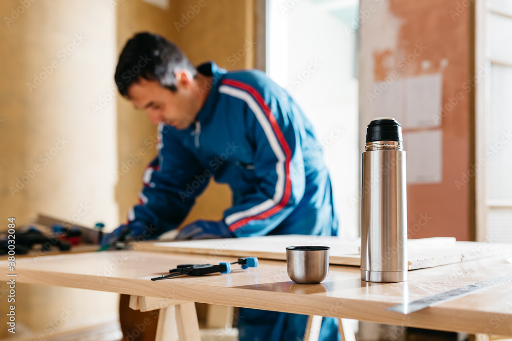 Metallic thermos on table against working carpenter. Metallic thermos on table against working carpenter.