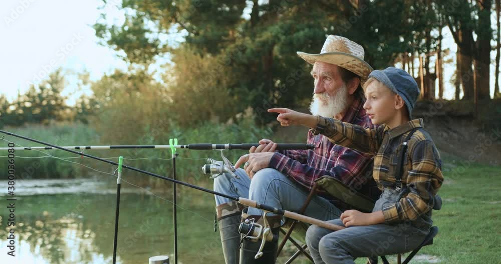 Side view of happy positive small boy which fishing together with his attractive respected bearded granddad on the pond and telling about his dream of big fish Side view of happy positive small boy which fishing together with his attractive respected bearded granddad on the pond and telling about his dream of big fish