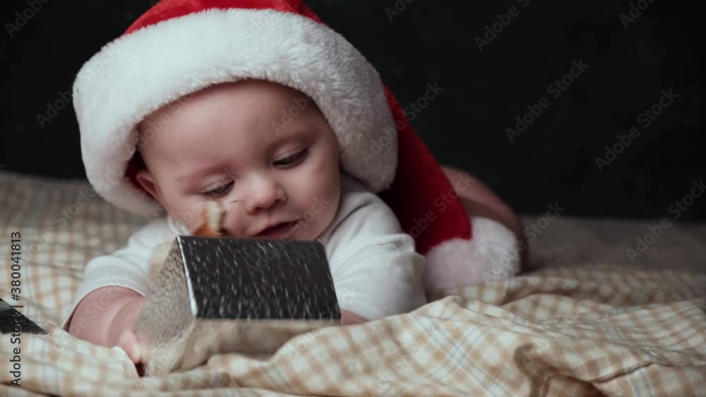 Funny little baby holding Christmas gift with toy bull - symbol of the 2021 year. Cute infant boy wearing Santa hat lying on sofa.