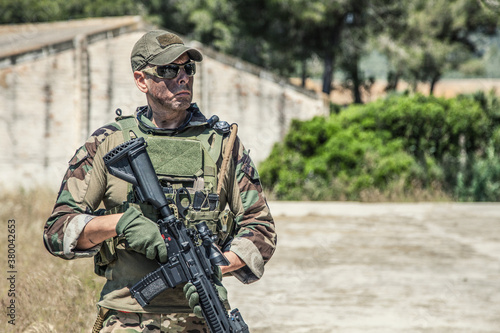 Navy SEALs fighter in ballistic goggles, equipped military ammunition and body armour, holding service rifle, looking in camera while standing outdoors. Special forces soldier half-length portrait