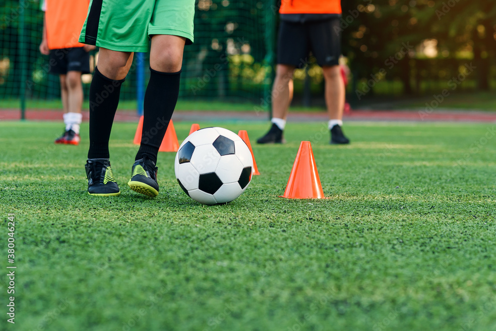 Persistent teen boy in sportswear trains football on soccer field and learns to circle the ball between training cones.