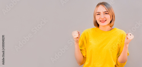 Photo of cheerful beautiful young woman standing isolated over gray wall background. Looking camera showing winner gesture.
