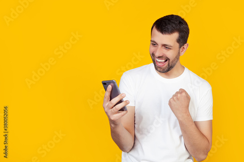 Young man with a beard in a white t-shirt shouts with pride and celebrates victory and success with a smartphone, very excited, jubilant emotion. Stands on isolated yellow background © Yauhen