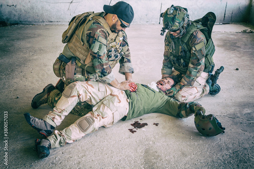 Photography Soldiers trying to stop bleeding at wounded comrade who lying on floor, suffering and screaming in paine