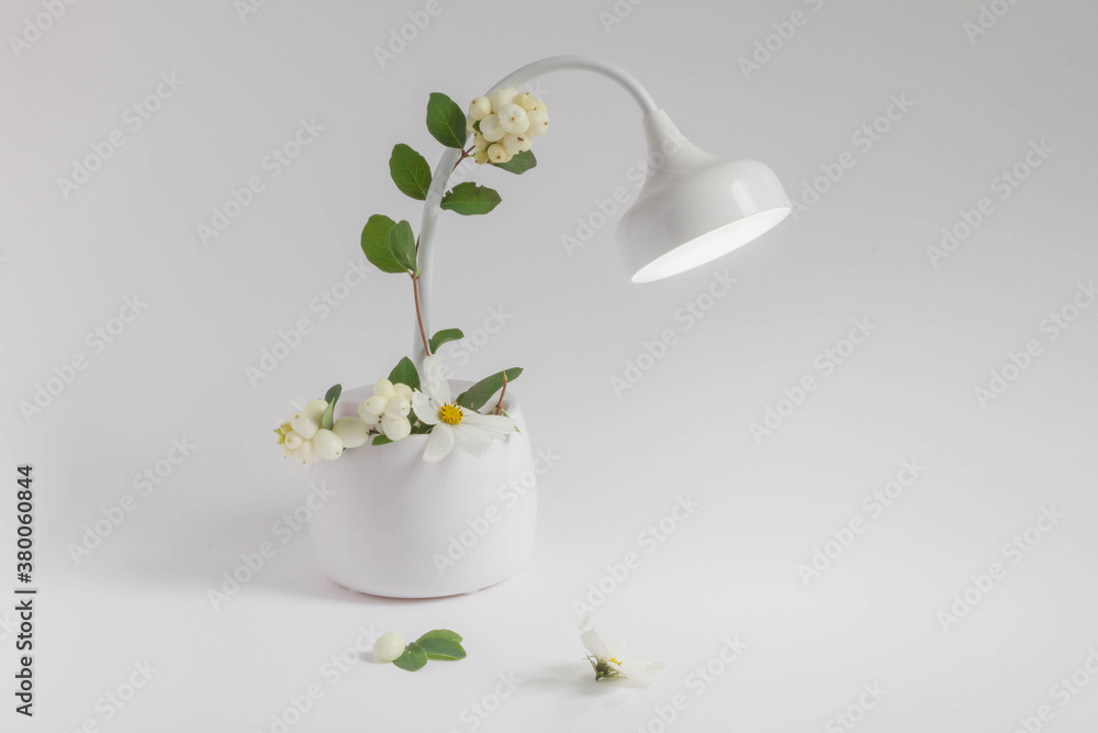 White flowers for decorating a white table lamp. White-green background of the workspace. Beautiful white Desk for office or home. White flowers for decorating a white table lamp. White-green background of the workspace. Beautiful white Desk for office or home.