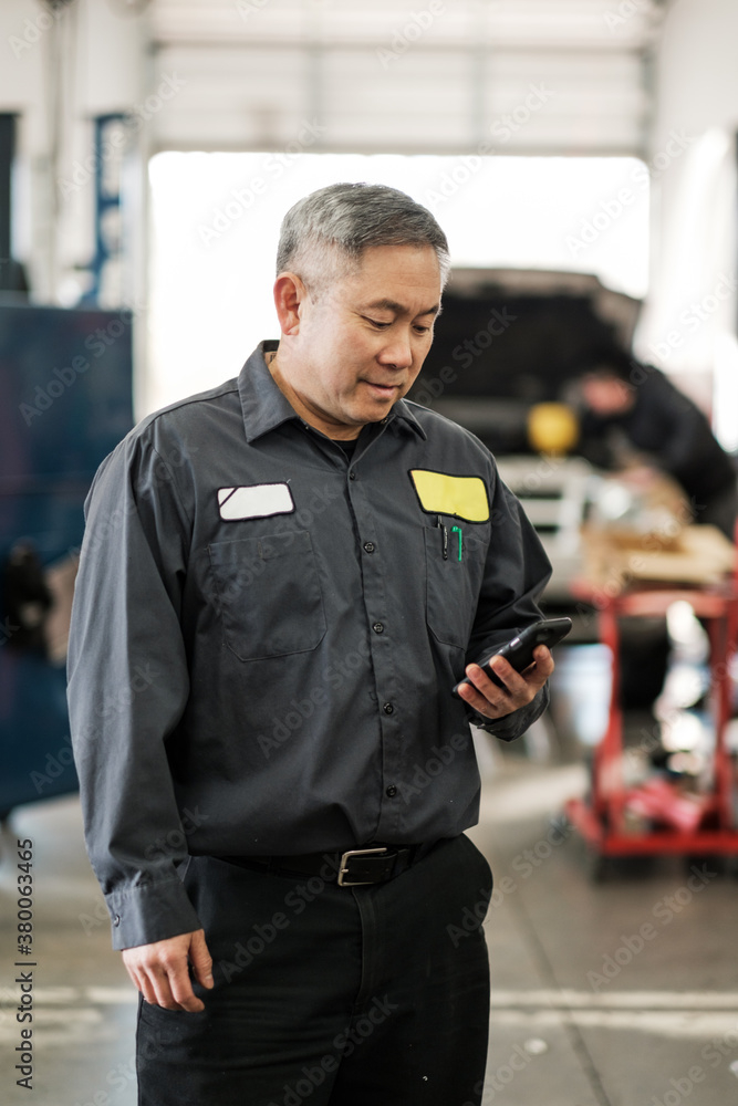 Asian car mechanic checking his phone in his auto repair shop Asian car mechanic checking his phone in his auto repair shop