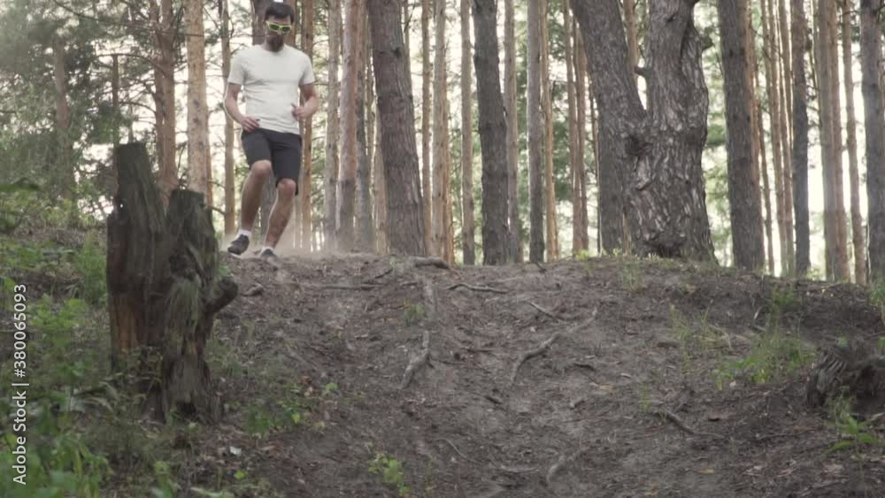 Slow motion Caucasian male athlete runs on a hill in the forest at sunset. A male runner is engaged in cross country jogging on a mountain rough terrain road. Trail running for beginner athletes. Slow motion Caucasian male athlete runs on a hill in the forest at sunset. A male runner is engaged in cross country jogging on a mountain rough terrain road. Trail running for beginner athletes.