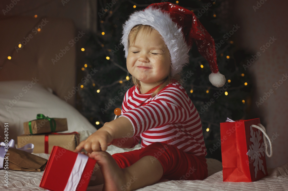 Happy Holidays! Adorable baby girl under Christmas tree with gift boxes. Christmas present. Cure girl in Santa hat open gift box. Happy Holidays! Adorable baby girl under Christmas tree with gift boxes. Christmas present. Cure girl in Santa hat open gift box.