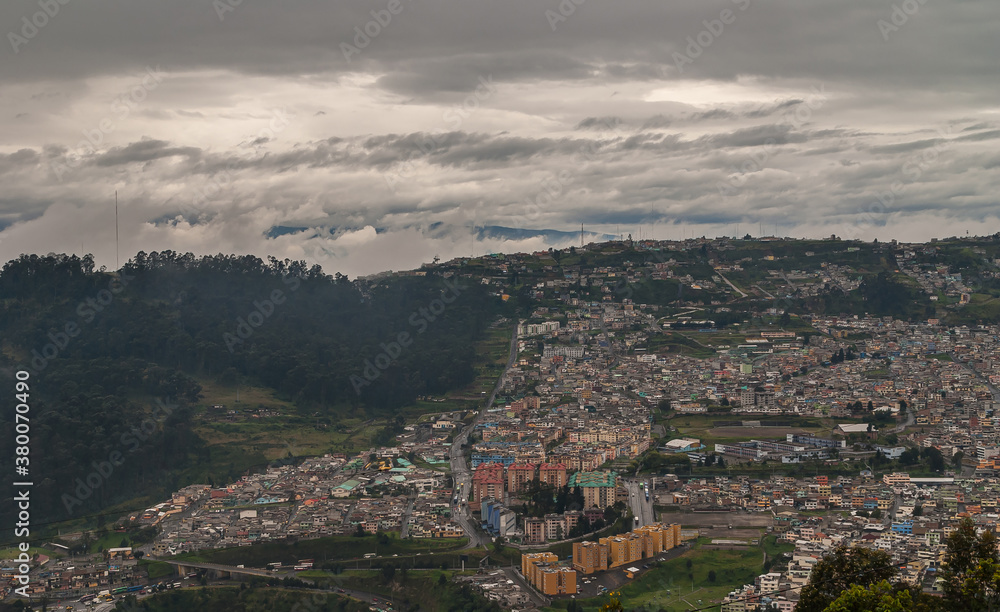 Quito, Ecuador - December 2, 2008: Seen from Cerro El Panecillo down. Wide cityscape with towers and regular housing under intense brown and white cloudscape with forested hill on side.
