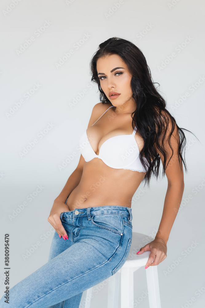 sexy young woman in bra and blue jeans sitting on stool and looking at  camera on grey Stock Photo | Adobe Stock
