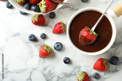 Dipping strawberry into fondue pot with chocolate on white marble table, flat lay