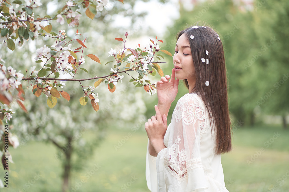 Beautiful Asian woman in a white dress, standing near a tree with white flowers and holding branches with her hands, looking away
