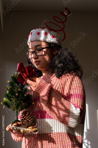 Curly latin girl wearing a red christmas hat and christmas sweater at home with a little christmas tree 