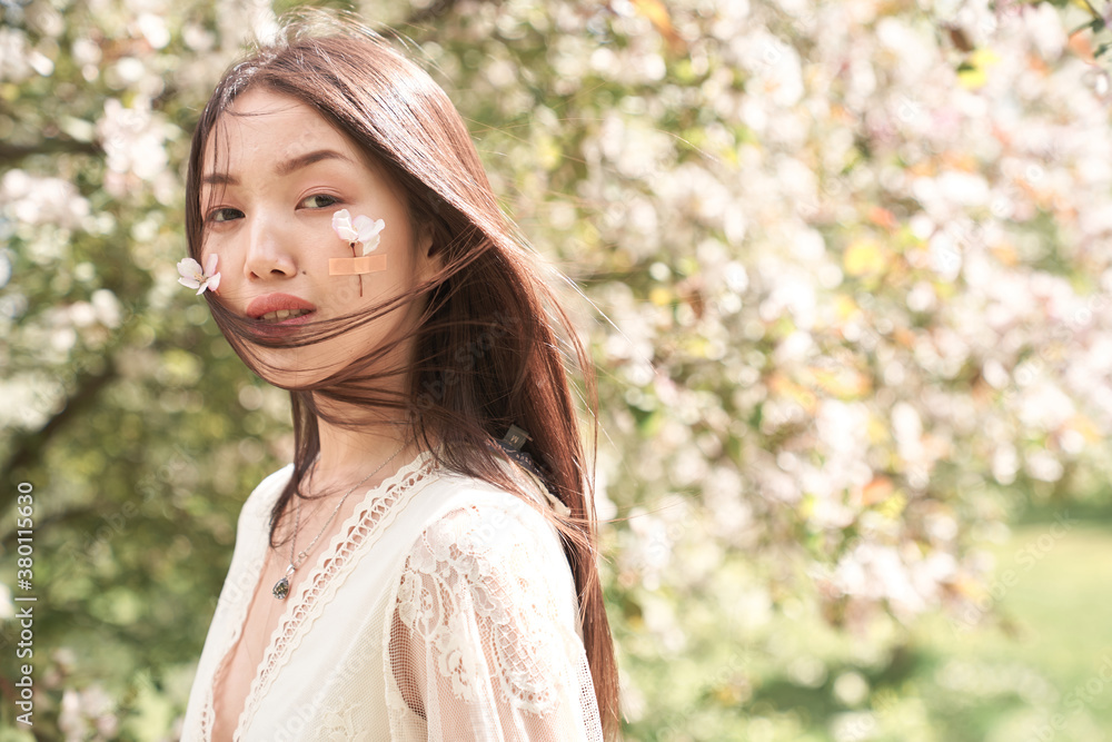 Portrait of a beautiful Asian woman in a white dress, she is standing near a tree with white flowers, on her cheeks flowers with petals are glued on a band-aid Portrait of a beautiful Asian woman in a white dress, she is standing near a tree with white flowers, on her cheeks flowers with petals are glued on a band-aid