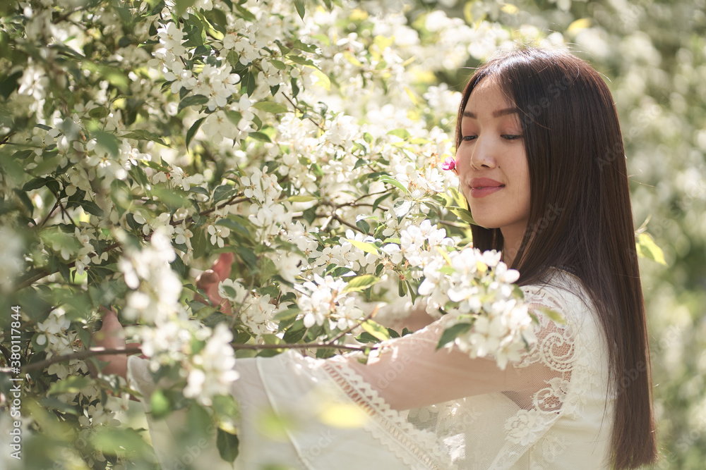 Beautiful Asian woman in a white dress, standing near a tree with white flowers and holding branches with her hands, looking away Beautiful Asian woman in a white dress, standing near a tree with white flowers and holding branches with her hands, looking away