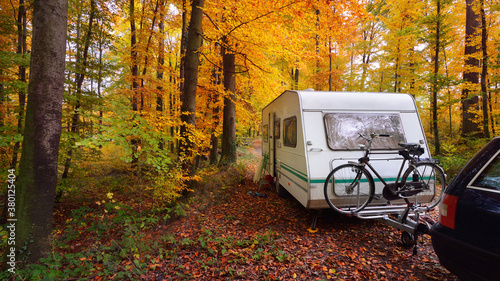 Caravan trailer with a bicycle and a car parked in a golden beech tree forest. Colorful red, orange and yellow leaves on the ground. Autumn landscape. Leisure activity in Heidelberg, Germany