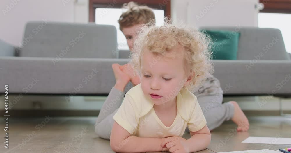Close up of a little girl, who is on the floor. Girl is looking at her drawing, brother jumps behind her and sits on the floor smiling. Grey sofa in the background. Close up of a little girl, who is on the floor. Girl is looking at her drawing, brother jumps behind her and sits on the floor smiling. Grey sofa in the background.