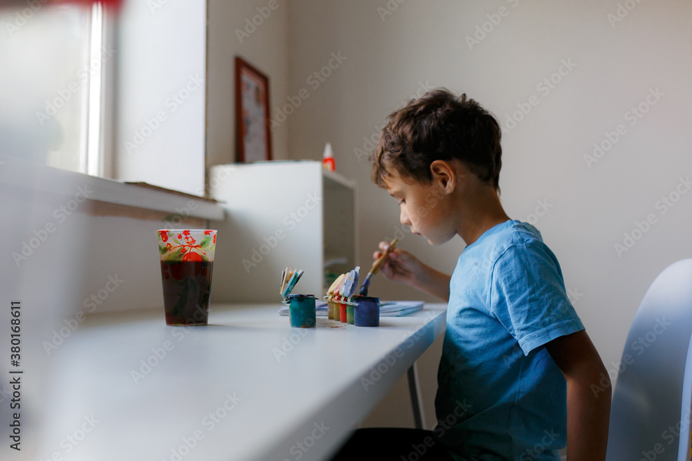 Curly dark-haired boy in a blue T-shirt draws a picture with paints while sitting at a white table Curly dark-haired boy in a blue T-shirt draws a picture with paints while sitting at a white table
