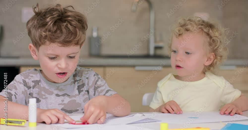 Small boy puts the glue on the cutted from paper objects, during making applications with coloured paper and glue, while sitting with his sister at the table in the kitchen. His sister looks at his Small boy puts the glue on the cutted from paper objects, during making applications with coloured paper and glue, while sitting with his sister at the table in the kitchen. His sister looks at his