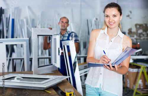 Young cheerful positive smiling woman manager standing with clipboard in workshop for production of plastic windows