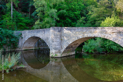 An ancient stone bridge mirroring in a small creek with trees and bushes in the background