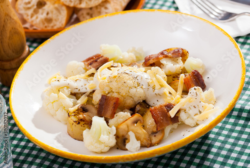 Appetizing baked potatoes served with cauliflower, bacon and mushrooms with sauce and grated cheese on white plate..