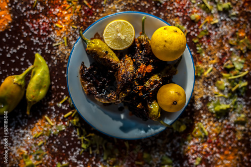Close up shot of Classic Bharela Marcha or Stuffed fried chilies in a glass plate along with lemons and all the spices needed for preparing it on a brown wooden surface. photo
