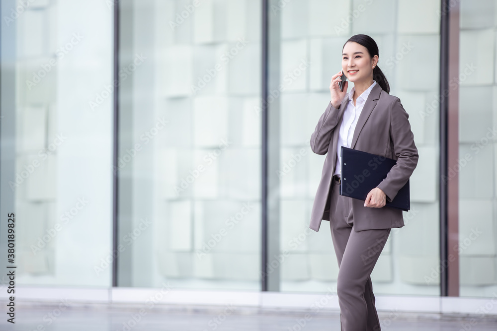 Asian woman with smartphone walking against street blurred building background, Fashion business photo of beautiful girl in casual suite with smart phone. 
