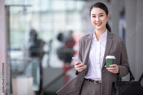 Asian woman with smartphone standing against street blurred building background. Fashion business photo of beautiful girl in casual suite with phone and cup of coffee photo