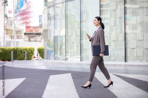 Asian woman with smartphone walking against street blurred building background, Fashion business photo of beautiful girl in casual suite with smart phone. 