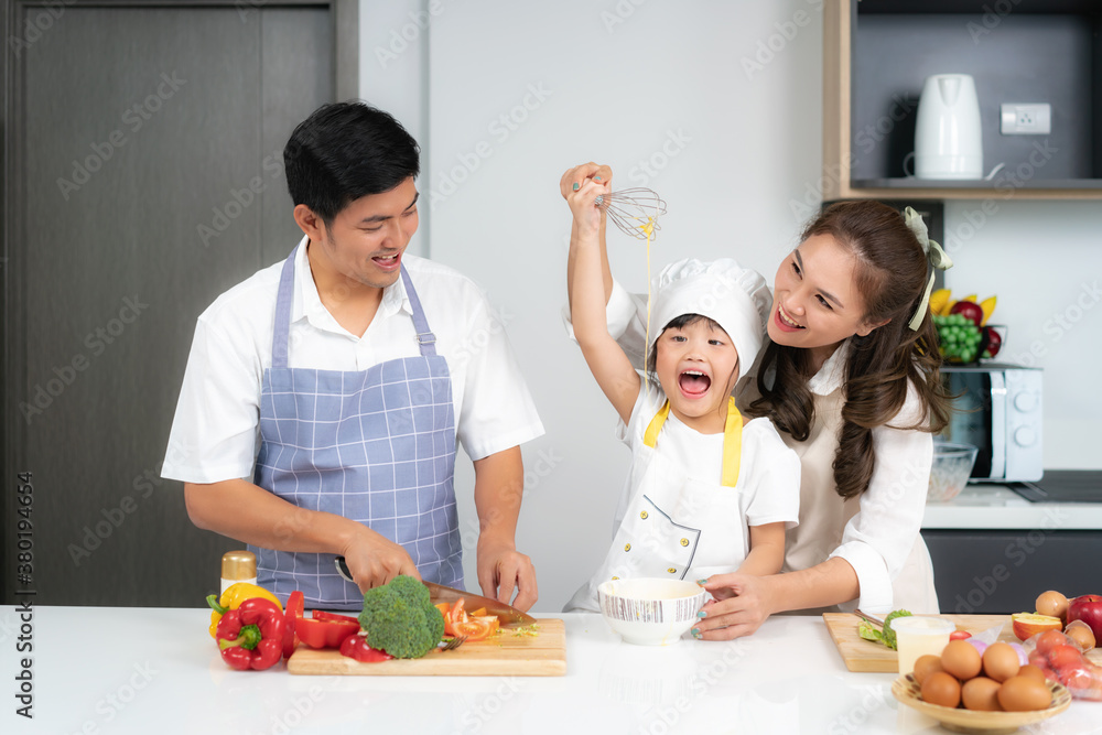 Asian family breaking an egg into the bowl and daughter whisking eggs in bowl on white table and looking happy to cooking food with her father and mother in kitchen at home.. Asian family breaking an egg into the bowl and daughter whisking eggs in bowl on white table and looking happy to cooking food with her father and mother in kitchen at home..
