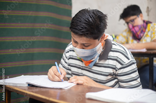 Indian boys students studying In classroom wearing mask maintaining social distancing looks at camera, school reopen during covid19 pandemic, new normal. selective focus
