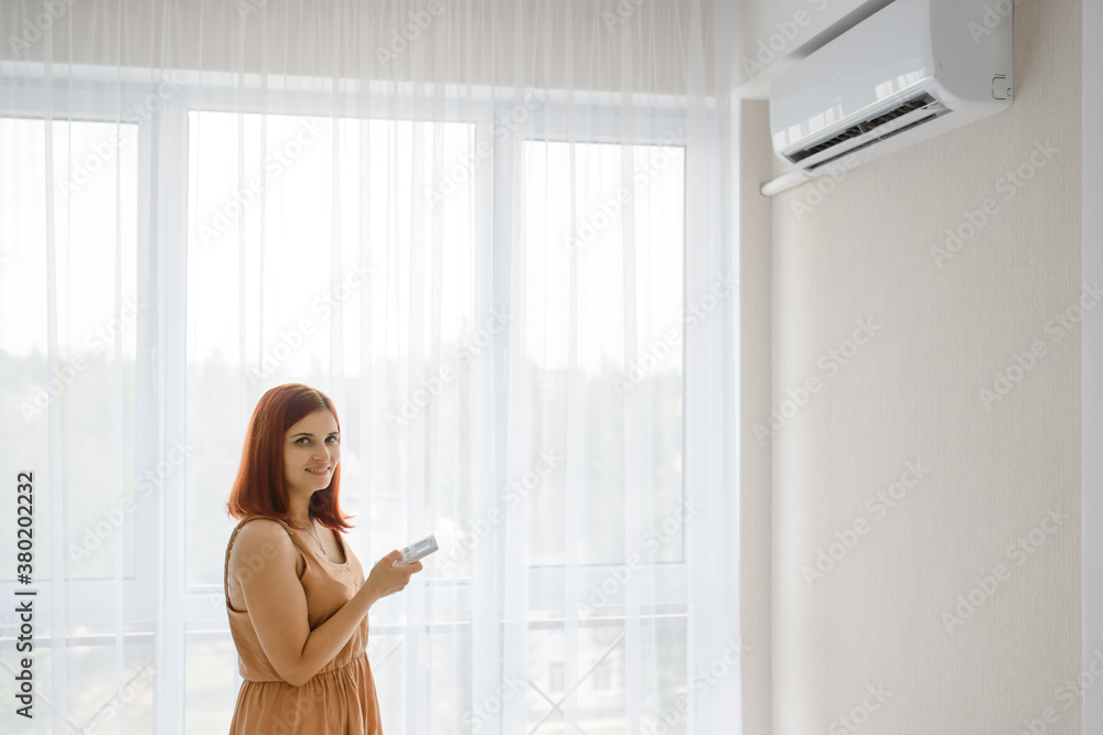 Young beautiful woman in her white apartment turns on the air conditioner and enjoys the cool.