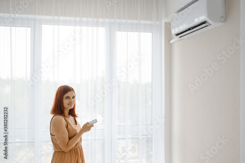 Young beautiful woman in her white apartment turns on the air conditioner and enjoys the cool.