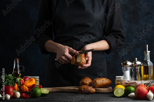 The chef in black apron cleans potatoes's skin on the professional restaurant's kitchen isolated on black background. Cooking tasty dish. Food concept.