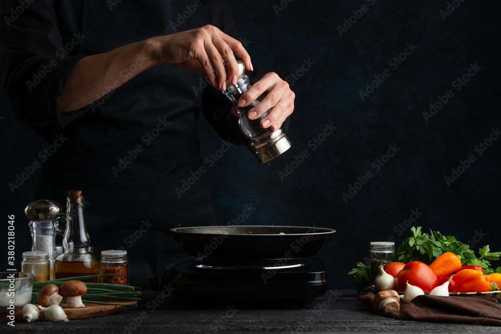 The chef in black adding the black pepper with grinder to the meal in frying pan isolated on dark background. Preparing delicious food with spices. For menu restaurant or recipe text. The chef in black adding the black pepper with grinder to the meal in frying pan isolated on dark background. Preparing delicious food with spices. For menu restaurant or recipe text.