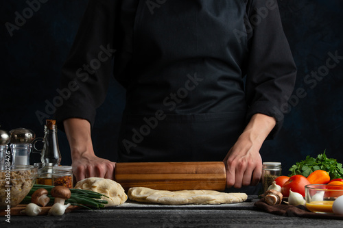 The chef in black with wooden rolling pin baking dough isolated on black background. Cooking pie. Traditional healthy food.