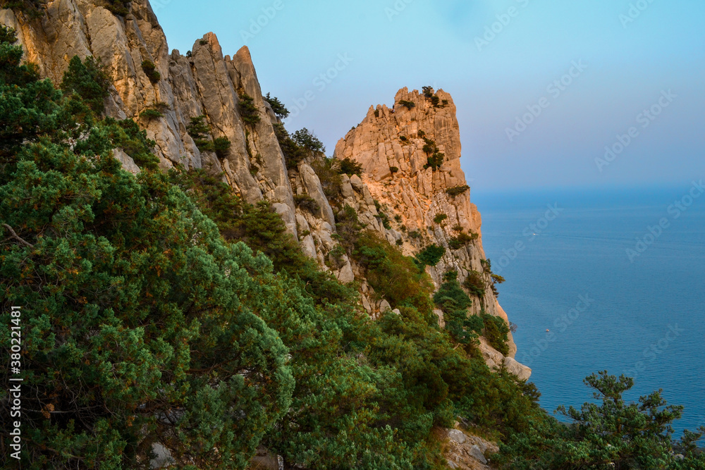 large sharp stones, rocks, cliff among green trees, bushes, forests in the warm orange light of the setting sun. Crimea. Summer landscape. Black sea backdrop, blue sky
