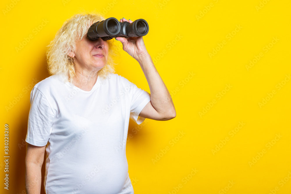 Old woman with a smile in a white t-shirt looks through binoculars on a yellow background Old woman with a smile in a white t-shirt looks through binoculars on a yellow background