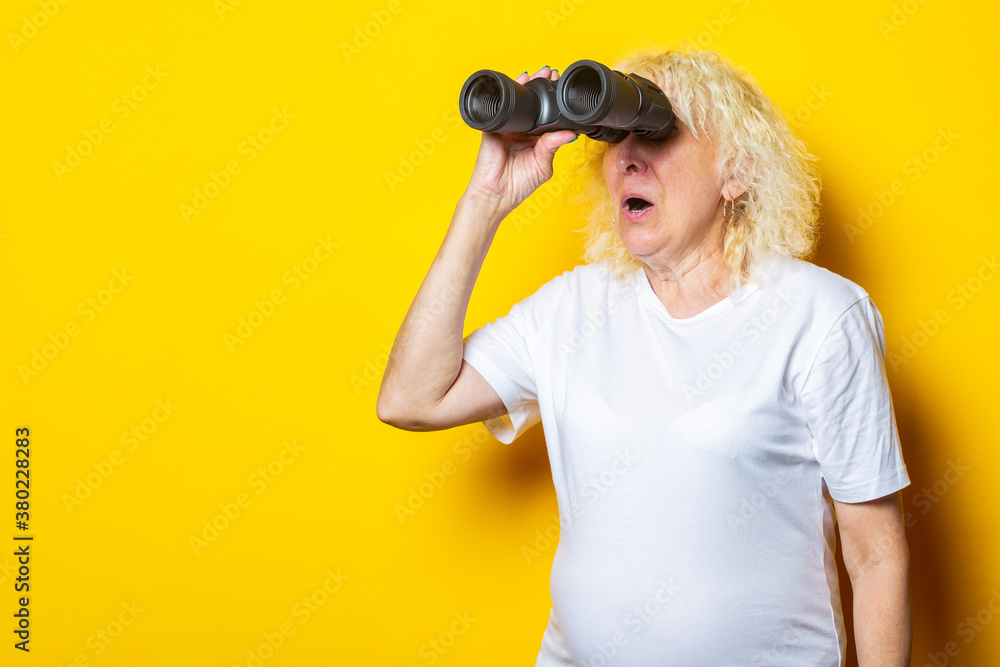 Old woman with a surprised face in a white t-shirt looks through binoculars on a yellow background Old woman with a surprised face in a white t-shirt looks through binoculars on a yellow background