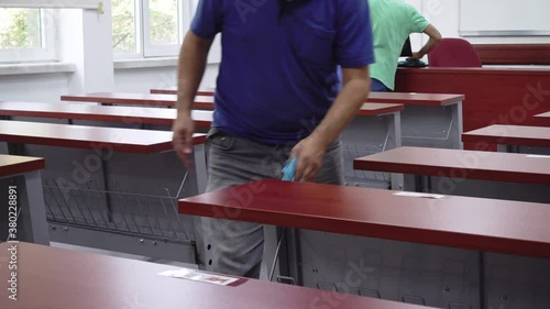 Two school janitors cleaning wooden desks of a empty university classroom