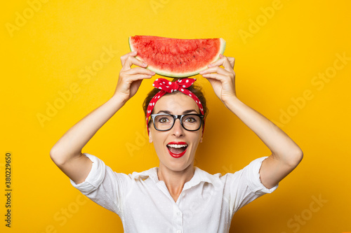 Young woman with surprised face with red headband and glasses holds watermelon above her head on yellow background