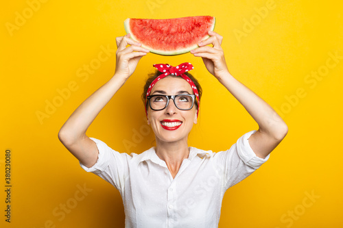 Young woman with a smile in a red headband and glasses holds a watermelon above her head on a yellow background