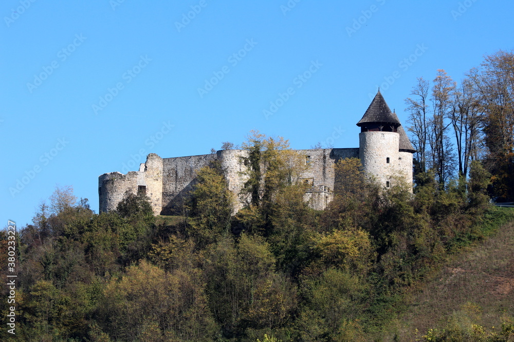 Abandoned old stone medieval town castle with two partially renovated guard towers on top of small hill surrounded with dense forest on clear blue sky background
