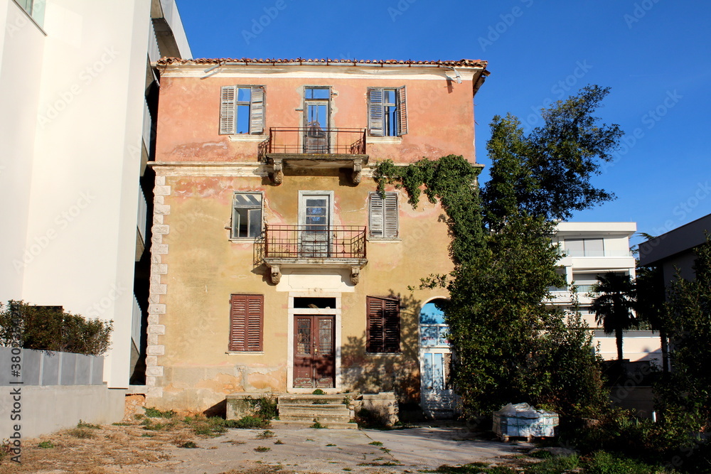 Creepy old abandoned apartment building with broken windows on dilapidated wooden frames and cracked facade surrounded with trees and new buildings on clear blue sky background
