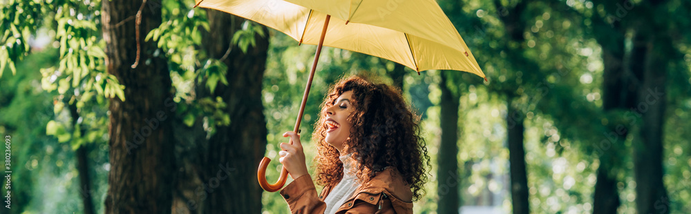 Panoramic shot of curly woman laughing while holding yellow umbrella in park - obrazy, fototapety, plakaty 