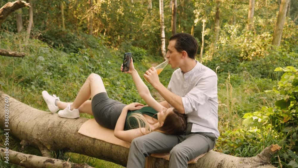 Young people couple date spend time together using phone take selfie pictures for social media drink bottled sweet beverage sitting on fallen tree trunk on nature countryside Young people couple date spend time together using phone take selfie pictures for social media drink bottled sweet beverage sitting on fallen tree trunk on nature countryside