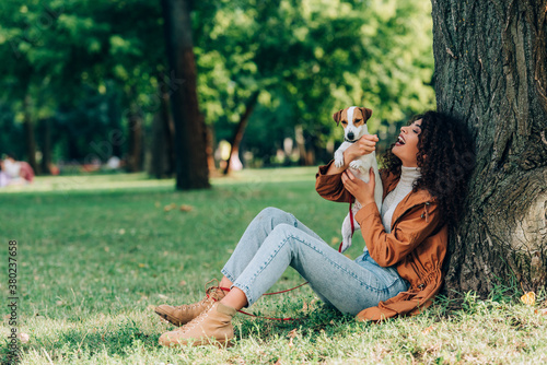 Selective focus of curly woman in jeans and raincoat holding jack russell terrier near tree in park