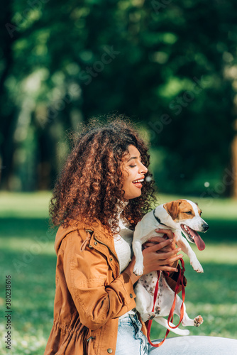 Curly woman in raincoat holding jack russell terrier with leash in park