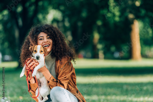 Young woman in raincoat holding jack russell terrier and looking at camera in park